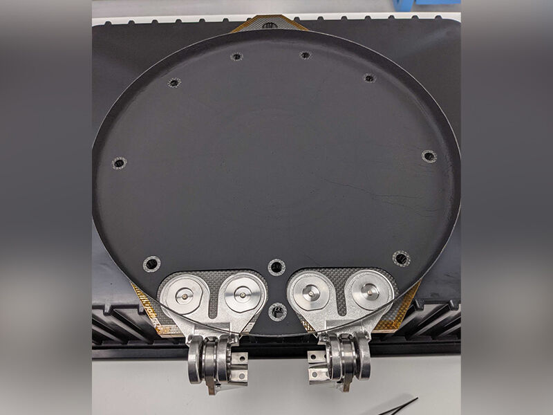 Image shows the underside of the Psyche deployable cover. The cover's interior is circular and smooth black with a curved edge. There are two hinge joins attached to the exterior carbon fiber sandwich panel exterior, which is just showing beneath edge of th ethe smooth surface.